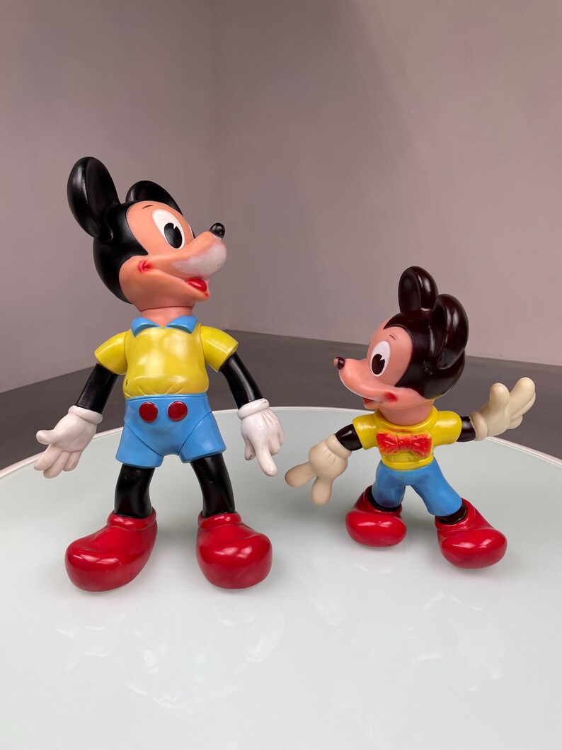 May include: Two plastic Mickey Mouse figurines. The larger figurine is wearing a yellow shirt, blue shorts, and red shoes. The smaller figurine is wearing a yellow shirt with a bow tie, blue pants, and red shoes.