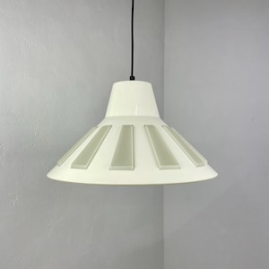 May include: A white pendant light with a wide, circular shade featuring a geometric design of vertical, rectangular cutouts. The light is suspended from a black cord.