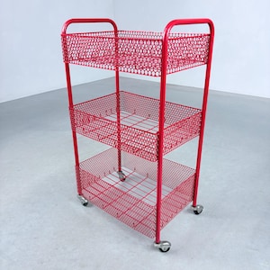 Può includere: Un carrello portaoggetti in metallo rosso brillante con tre cestini rettangolari in rete perforata. Il carrello presenta una struttura tubolare con una maniglia superiore ed è dotato di quattro piccole ruote piroettanti alla base.