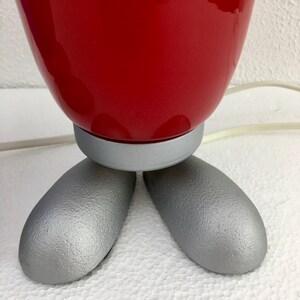 May include: A red ceramic table lamp with a silver base and three legs. The lamp is on a white surface.