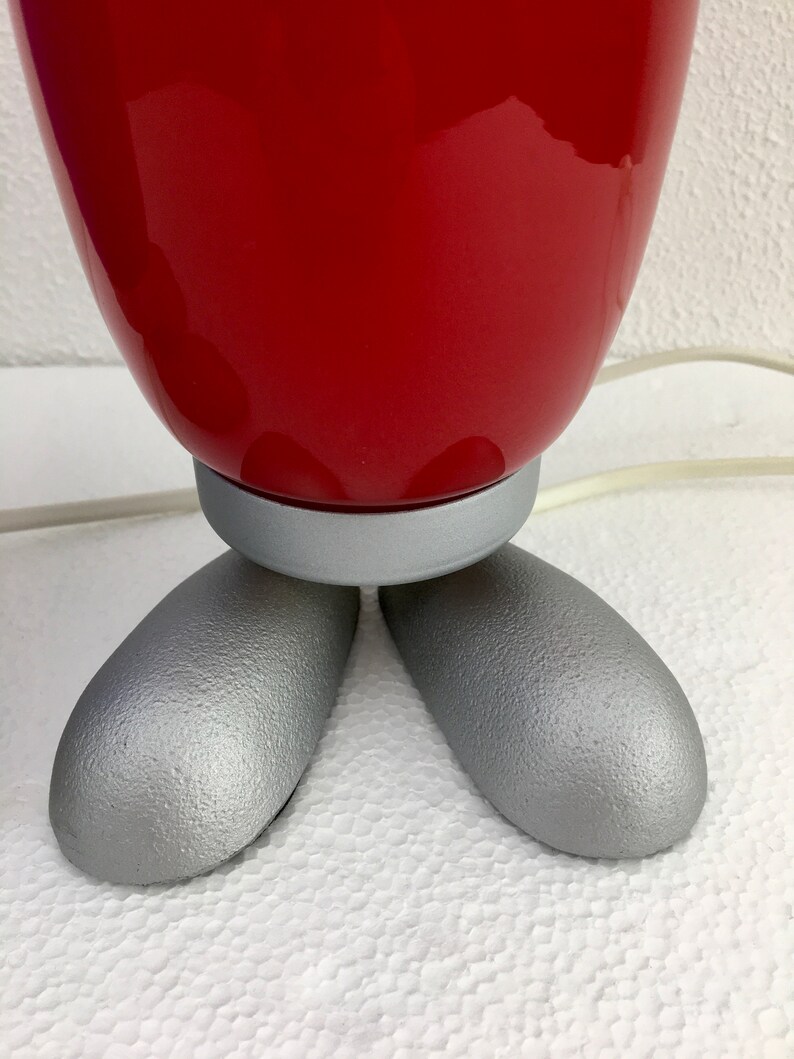 May include: A red ceramic table lamp with a silver base and three legs. The lamp is on a white surface.