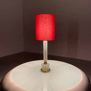 Op de afbeelding: Een rode lampenkap met een heldere glazen voet staat op een ronde witte tafel. De lampenkap is gemaakt van stof en heeft een getextureerd oppervlak. De voet is gemaakt van glas en heeft een gouden metalen accent.