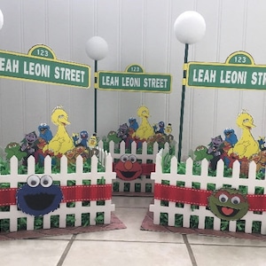 May include: Two Sesame Street themed centerpieces with white picket fences, green grass, and red ribbon. Each centerpiece features a street sign that reads "Leah Leoni Street 123" and a group of Sesame Street characters including Big Bird, Cookie Monster, Elmo, and Bert and Ernie.