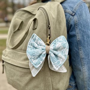 May include: A fabric bow keychain featuring a light blue and white floral pattern, accented with a tan center wrap. The bow is attached to a gold-toned metal lobster clasp, which is clipped onto the zipper pull of a solid olive green canvas backpack.