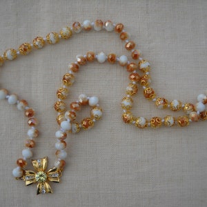 Può includere: Una collana di perline bianche e marroni con un ciondolo a forma di fiore dorato. Le perline sono sfaccettate e hanno un design a spirale.