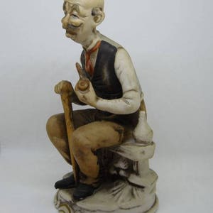 May include: A ceramic figurine of a man with a mustache and a pipe, sitting on a stone bench with a walking stick. He is wearing a brown vest and a white shirt.