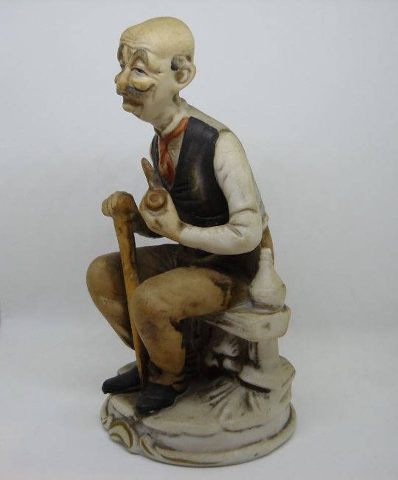May include: A ceramic figurine of a man with a mustache and a pipe, sitting on a stone bench with a walking stick. He is wearing a brown vest and a white shirt.