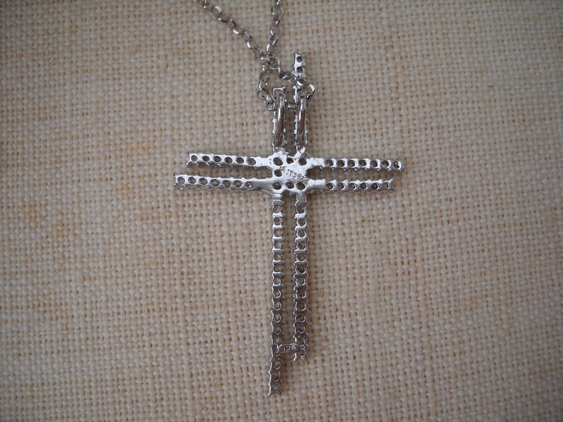 Pode incluir: Colar com pingente de cruz em tom prateado com uma dupla fileira de pedras transparentes. O pingente est&aacute; preso a uma corrente em tom prateado.