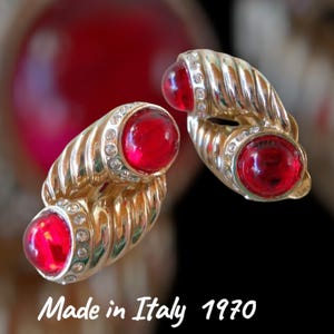 May include: Vintage gold-tone earrings with a twisted design, featuring large, round, red cabochon stones. The stones are surrounded by small, clear crystals. The text "Made in Italy 1970" is visible.