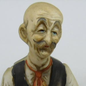 May include: A ceramic figurine of a man with a mustache and a painted-on smile. He is wearing a white shirt and a black vest with a red tie.