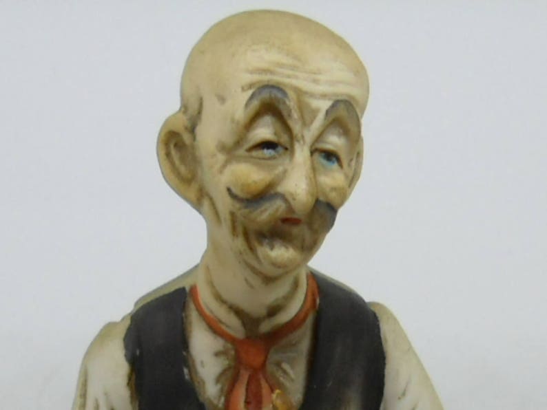 May include: A ceramic figurine of a man with a mustache and a painted-on smile. He is wearing a white shirt and a black vest with a red tie.