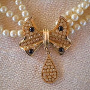 Op de afbeelding: Een vintage parelketting met een goudkleurige strik-hanger. De strik is bezet met heldere en zwarte strass-steentjes, en een druppelvormige bedel hangt eronder. De ketting heeft drie strengen imitatieparels.