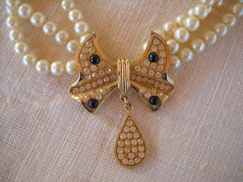 Op de afbeelding: Een vintage parelketting met een goudkleurige strik-hanger. De strik is bezet met heldere en zwarte strass-steentjes, en een druppelvormige bedel hangt eronder. De ketting heeft drie strengen imitatieparels.