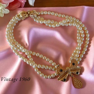 Op de afbeelding: Vintage jaren 60 parelketting met drie strengen en een goudkleurige strik-hanger. De hanger is versierd met strass en donkerblauwe accenten. De ketting ligt op een roze satijnen stof.