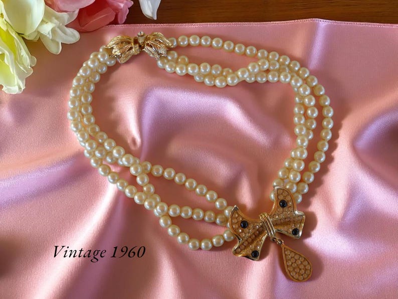 Op de afbeelding: Vintage jaren 60 parelketting met drie strengen en een goudkleurige strik-hanger. De hanger is versierd met strass en donkerblauwe accenten. De ketting ligt op een roze satijnen stof.