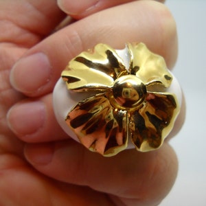 May include: A white ceramic ring with a gold flower design. The flower has a raised center and petals with a textured surface.