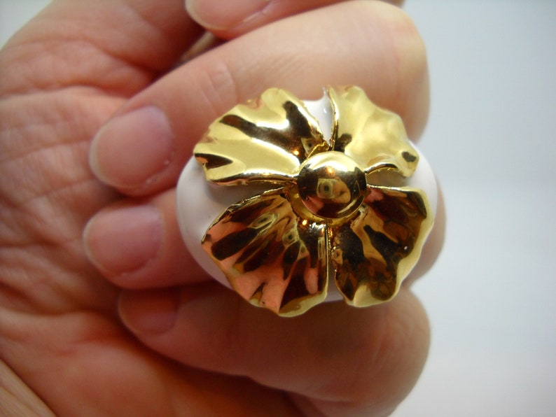 May include: A white ceramic ring with a gold flower design. The flower has a raised center and petals with a textured surface.