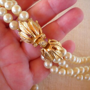 Op de afbeelding: Een vintage parelketting met een goudkleurige sluiting. De sluiting heeft een bladvorm met een centrale heldere strass-steen. De ketting heeft meerdere strengen ronde, cr&egrave;mekleurige parels.