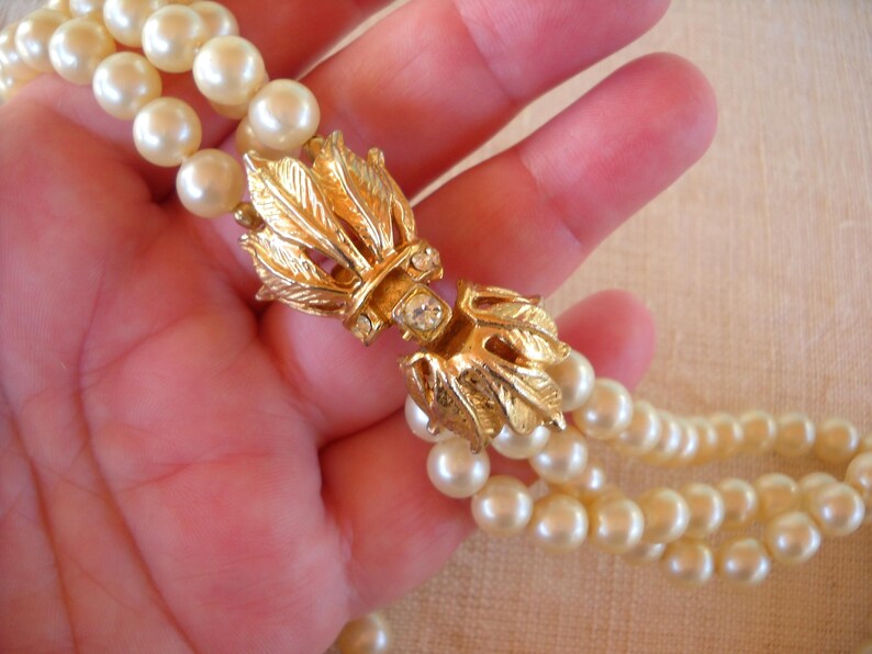 Op de afbeelding: Een vintage parelketting met een goudkleurige sluiting. De sluiting heeft een bladvorm met een centrale heldere strass-steen. De ketting heeft meerdere strengen ronde, cr&egrave;mekleurige parels.