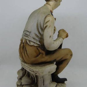 May include: A ceramic figurine of a man sitting on a stone bench. The man is wearing a white shirt and brown pants. The figurine is painted in shades of brown, white, and black.
