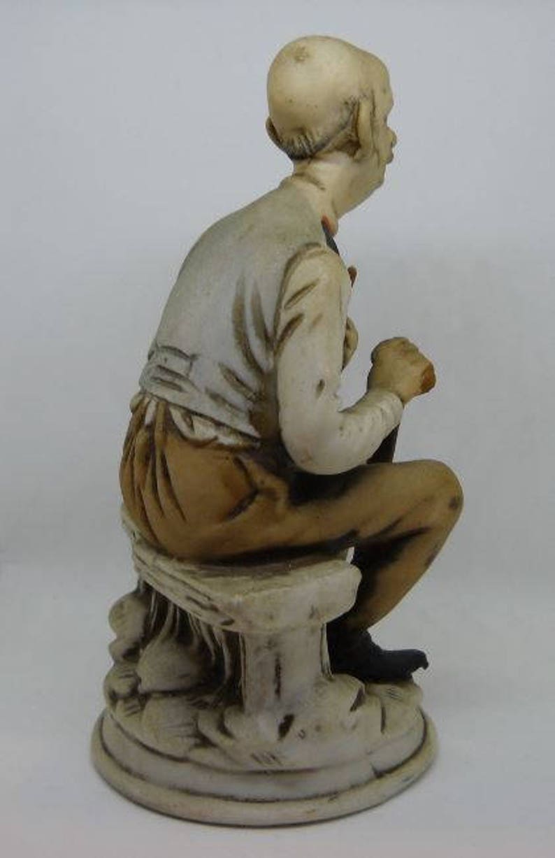 May include: A ceramic figurine of a man sitting on a stone bench. The man is wearing a white shirt and brown pants. The figurine is painted in shades of brown, white, and black.