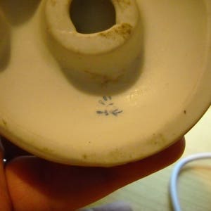 May include: Close-up of the bottom of a white porcelain figurine. The bottom has a blue mark with the letters "D-E" inside.