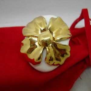 May include: A white porcelain ring holder with a gold flower design. The ring holder is sitting on a red velvet pouch.