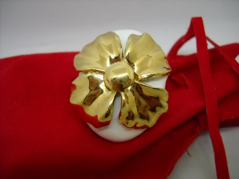 May include: A white porcelain ring holder with a gold flower design. The ring holder is sitting on a red velvet pouch.