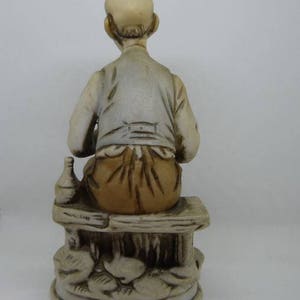 May include: A ceramic figurine of a man sitting on a bench, holding a bottle. The man is wearing a brown shirt and pants. The bench is decorated with floral details.