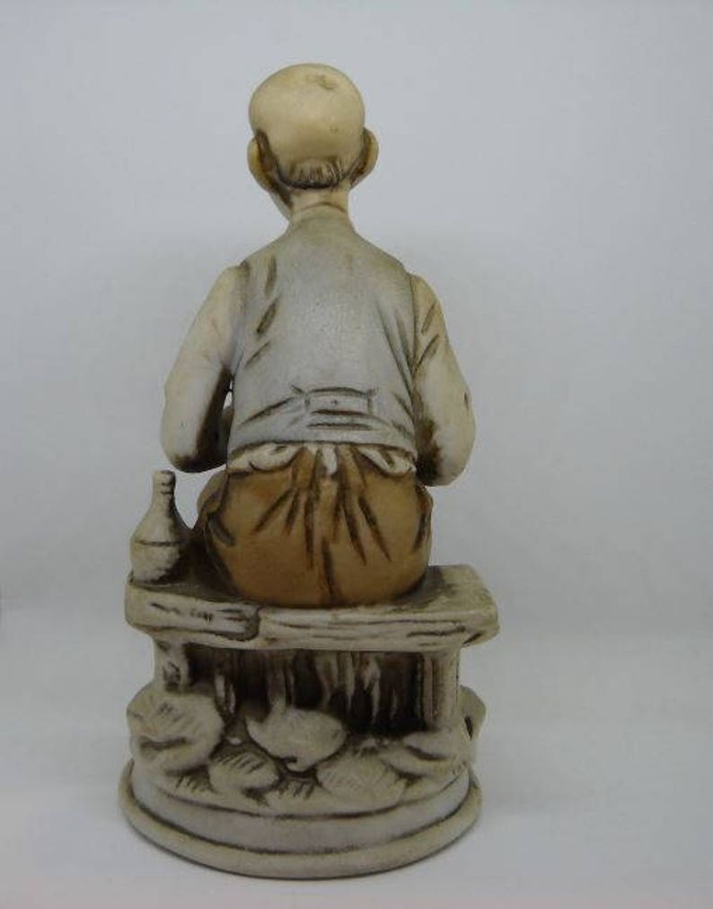 May include: A ceramic figurine of a man sitting on a bench, holding a bottle. The man is wearing a brown shirt and pants. The bench is decorated with floral details.