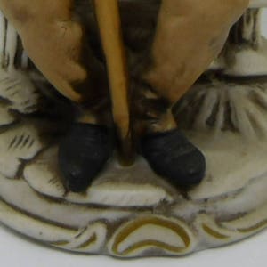 May include: A ceramic figurine of a person sitting on a base with a stick in their hand. The figurine is painted in shades of brown, black, and white.