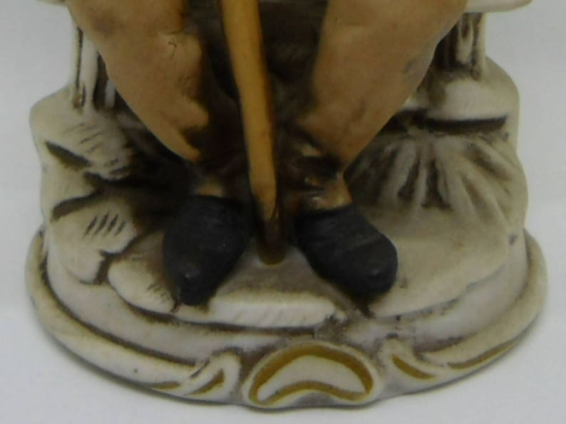 May include: A ceramic figurine of a person sitting on a base with a stick in their hand. The figurine is painted in shades of brown, black, and white.