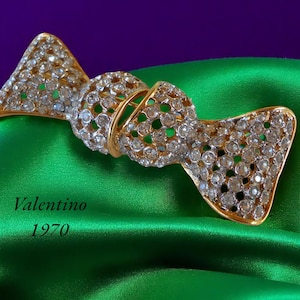 May include: A vintage Valentino bow brooch from 1970, featuring a gold-tone metal frame encrusted with clear rhinestones. The brooch is set against a vibrant green satin fabric with a purple background.