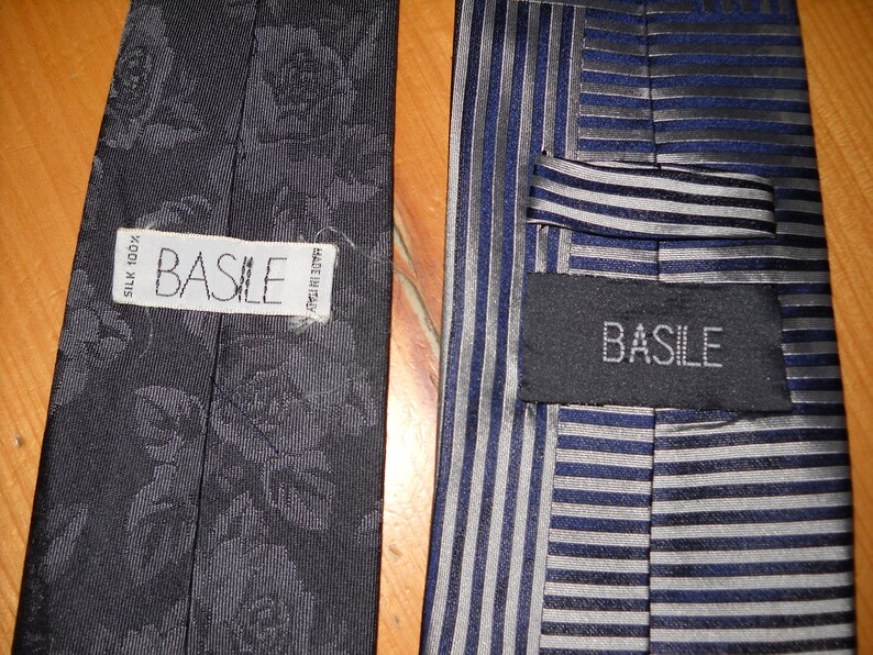 May include: Two black silk ties with the brand name "BASILE" printed on the label. One tie has a floral pattern and the other has a striped pattern.