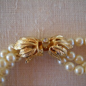 Op de afbeelding: Een parelketting met dubbele streng en een goudkleurige sluiting. De sluiting heeft de vorm van een strik met bladdetails en een kleine, heldere edelsteen in het midden. De parels zijn cr&egrave;mewit.