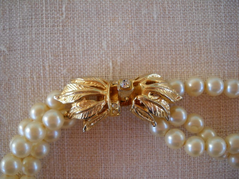 Op de afbeelding: Een parelketting met dubbele streng en een goudkleurige sluiting. De sluiting heeft de vorm van een strik met bladdetails en een kleine, heldere edelsteen in het midden. De parels zijn cr&egrave;mewit.