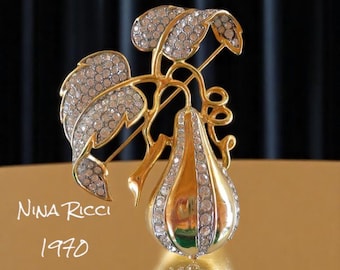 May include: A gold-tone brooch featuring a pear-shaped design with clear rhinestones. The pear is attached to a gold-tone stem with leaves that are also covered in rhinestones. The brooch is signed "Nina Ricci 1970".