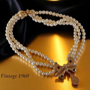 Op de afbeelding: Vintage parelketting uit de jaren 1960 met drie strengen imitatieparels. De ketting heeft een goudkleurige strik-hanger met heldere en donkerblauwe strass-steentjes. De sluiting is ook goudkleurig met blad details.