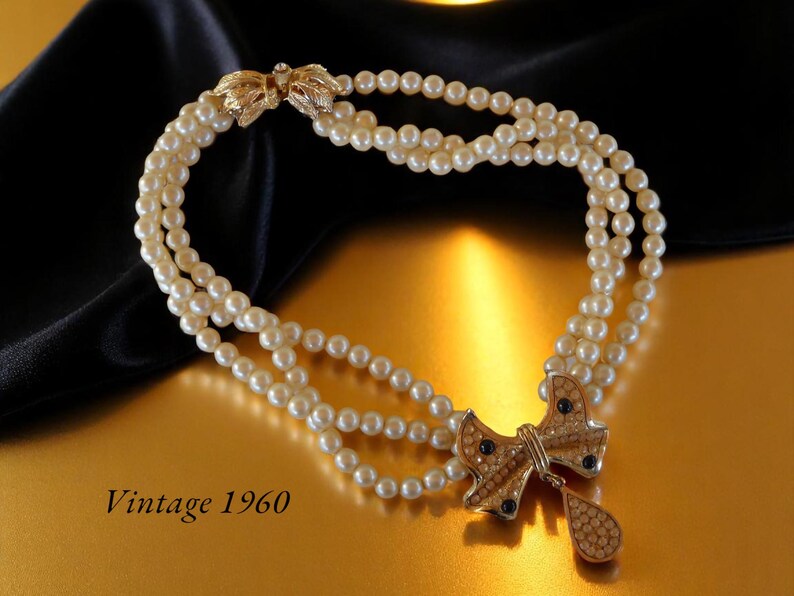 Op de afbeelding: Vintage parelketting uit de jaren 1960 met drie strengen imitatieparels. De ketting heeft een goudkleurige strik-hanger met heldere en donkerblauwe strass-steentjes. De sluiting is ook goudkleurig met blad details.