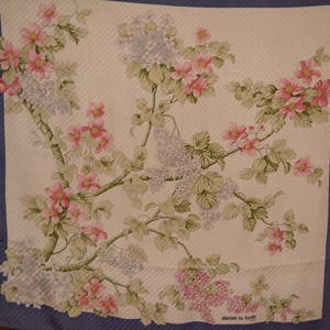 May include: A white silk scarf with a floral pattern of pink and purple flowers and green leaves. The scarf has a blue border and the text "daniel la fonti" in the bottom right corner.