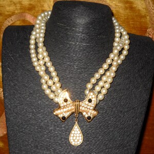 Op de afbeelding: Een parelketting met drie strengen en een goudkleurige strik-hanger. De strik is versierd met heldere strass-steentjes en zwarte accenten. Een druppelvormige hanger hangt eronder. De ketting wordt getoond op een zwarte mannequin.