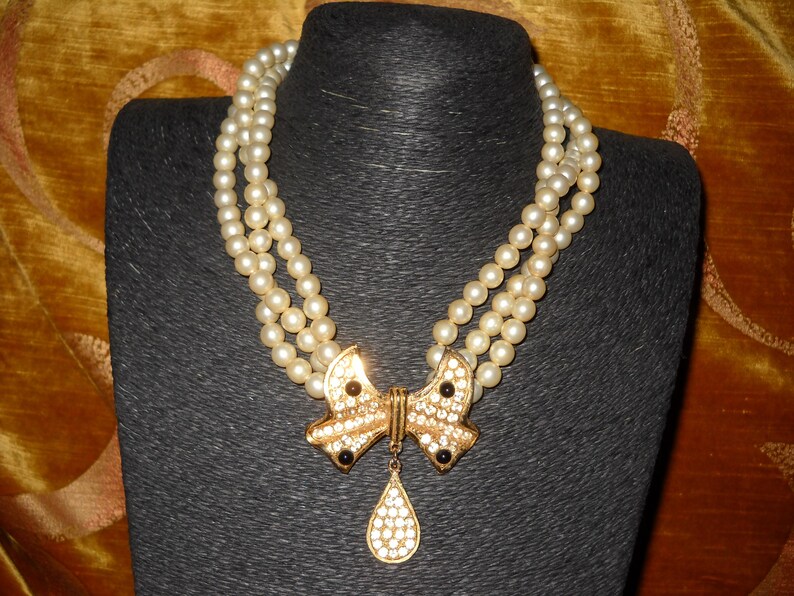 Op de afbeelding: Een parelketting met drie strengen en een goudkleurige strik-hanger. De strik is versierd met heldere strass-steentjes en zwarte accenten. Een druppelvormige hanger hangt eronder. De ketting wordt getoond op een zwarte mannequin.