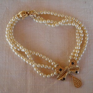 Op de afbeelding: Een parelketting met drie strengen en een goudkleurige strik-hanger. De strik-hanger heeft zwarte accenten en een druppel. De ketting wordt vastgemaakt met een gouden sluiting.