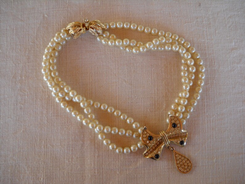 Op de afbeelding: Een parelketting met drie strengen en een goudkleurige strik-hanger. De strik-hanger heeft zwarte accenten en een druppel. De ketting wordt vastgemaakt met een gouden sluiting.