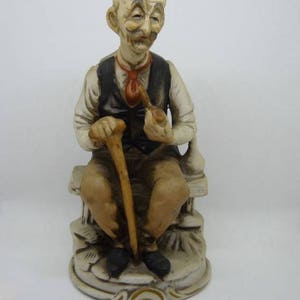 May include: A ceramic figurine of an elderly man sitting on a bench, holding a pipe and a walking stick. He is wearing a vest and a tie, and has a whimsical expression on his face.
