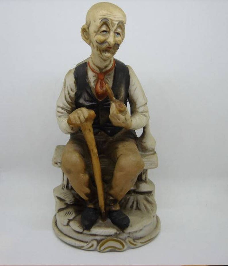 May include: A ceramic figurine of an elderly man sitting on a bench, holding a pipe and a walking stick. He is wearing a vest and a tie, and has a whimsical expression on his face.