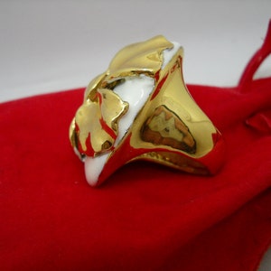 May include: A gold ring with a white ceramic design. The ring is shaped like a flower with a textured surface.