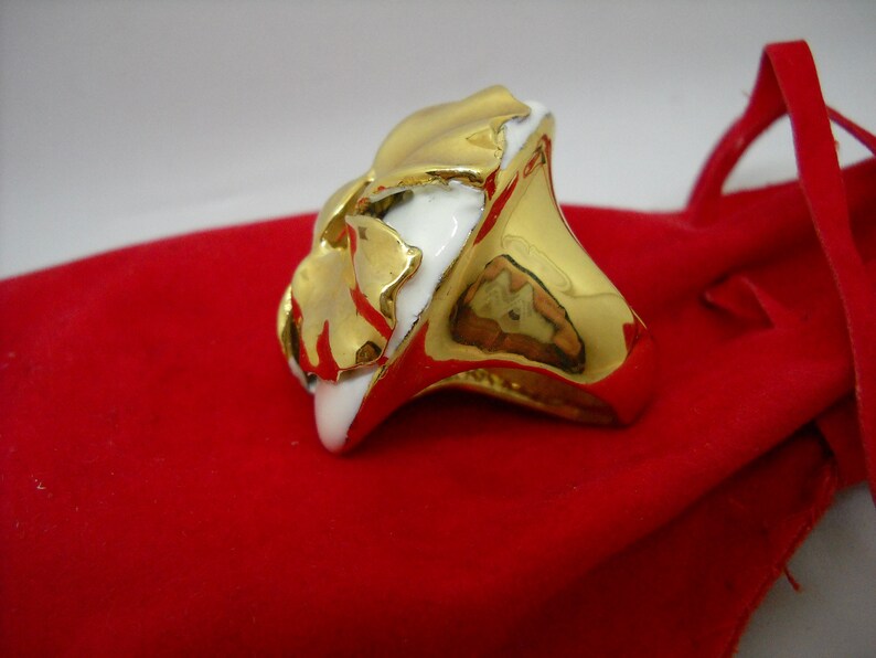 May include: A gold ring with a white ceramic design. The ring is shaped like a flower with a textured surface.