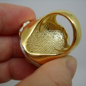 May include: Gold-tone ring with a white enamel design. The ring has a textured band and a large oval-shaped stone.
