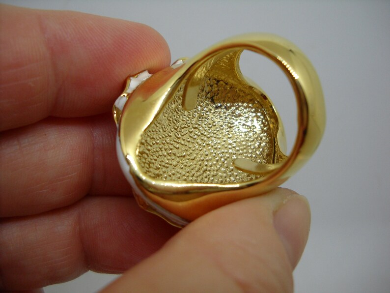 May include: Gold-tone ring with a white enamel design. The ring has a textured band and a large oval-shaped stone.